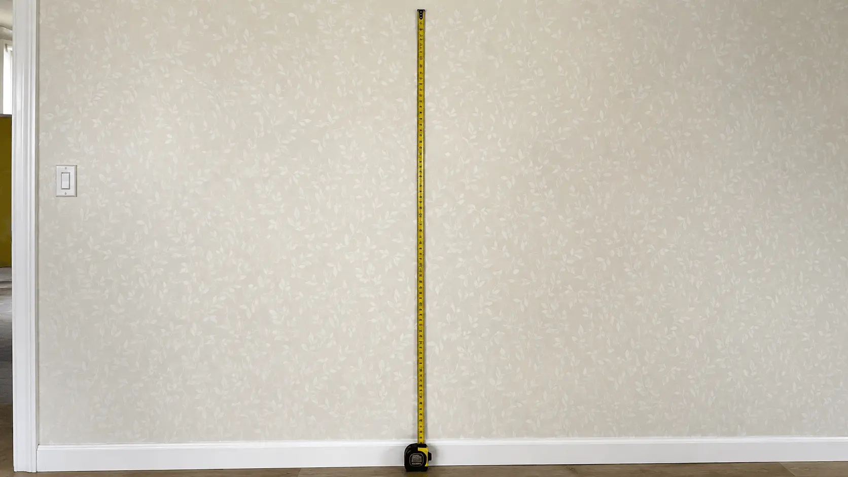Tape measure extended from skirting board to ceiling against a papered wall, next to a light switch